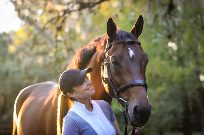 Libby Neily smiling in a cap beside a bay horse in a bridle under the oaks