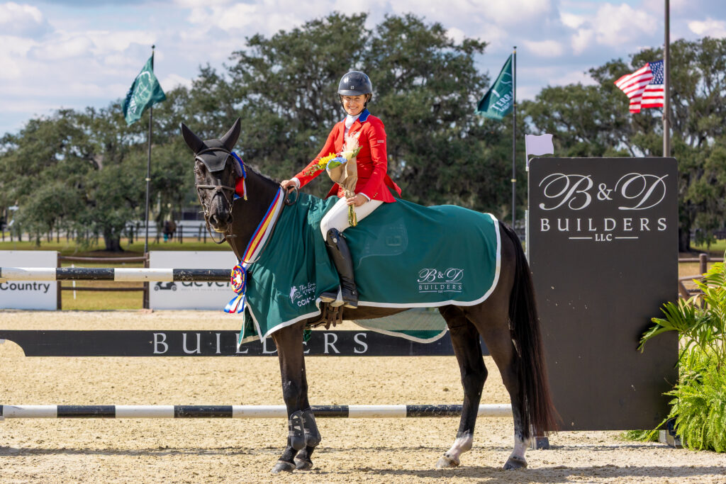 Rider in a red coat on a dark bay horse wearing a green cooler and tricolor ribbon