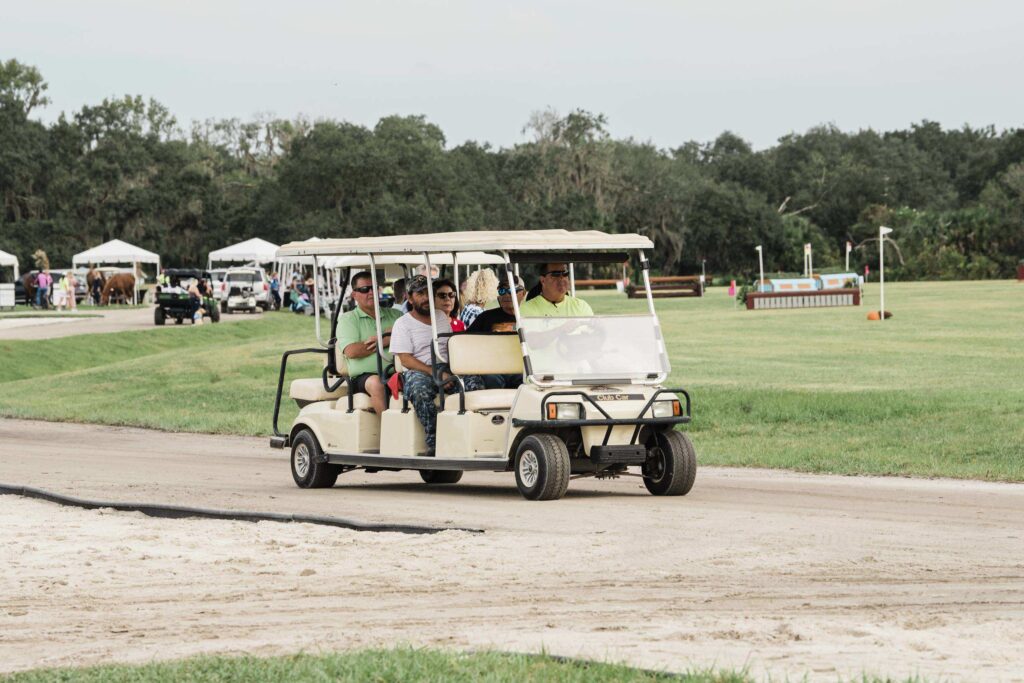 Staff drive guests in a long golf cart along a sand track past white tents