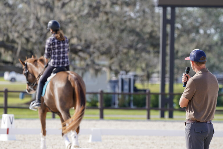 Clinician with a microphone watches a rider work a chestnut horse in the arena