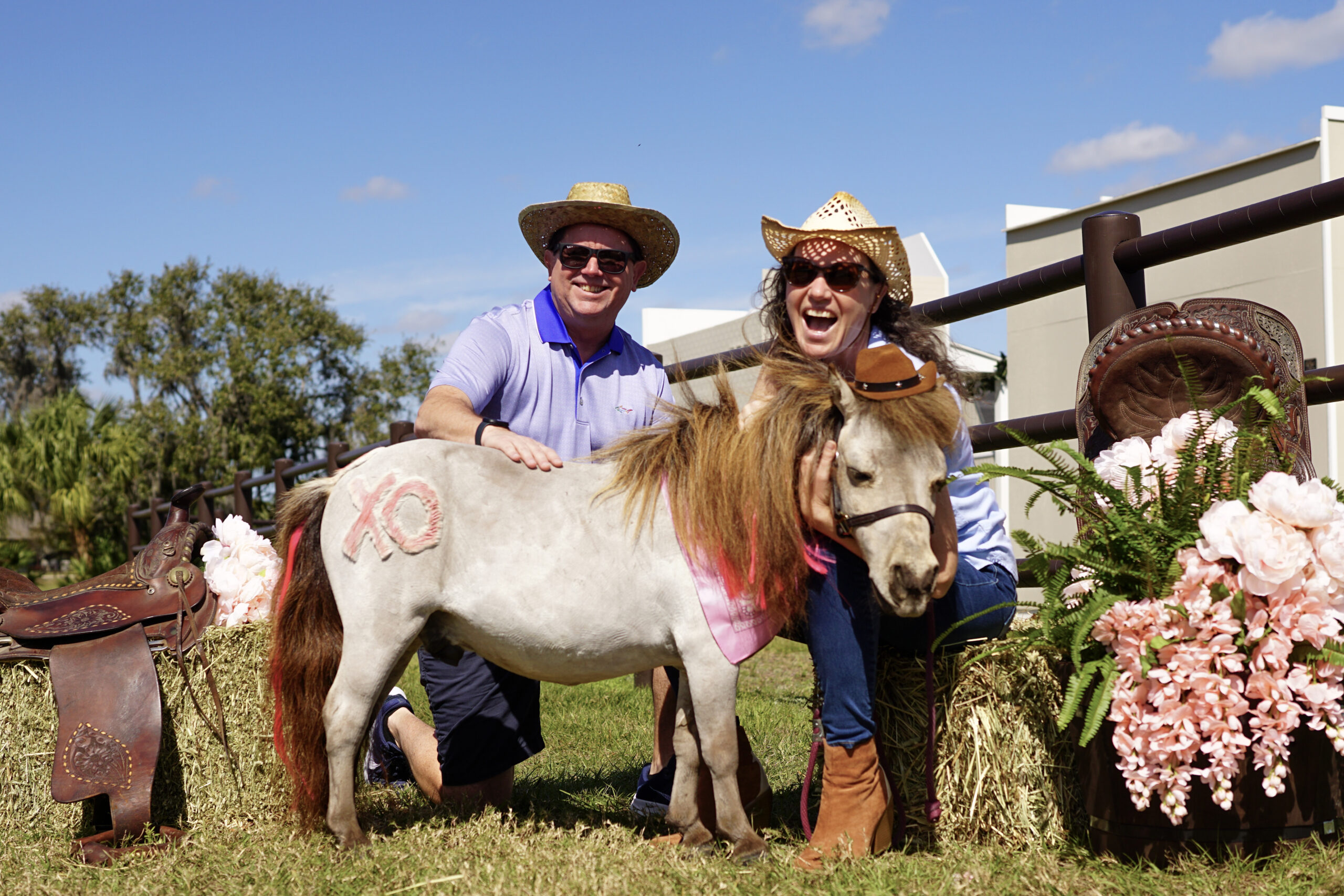 Two adults in straw hats kneel with a miniature horse painted with a pink XO