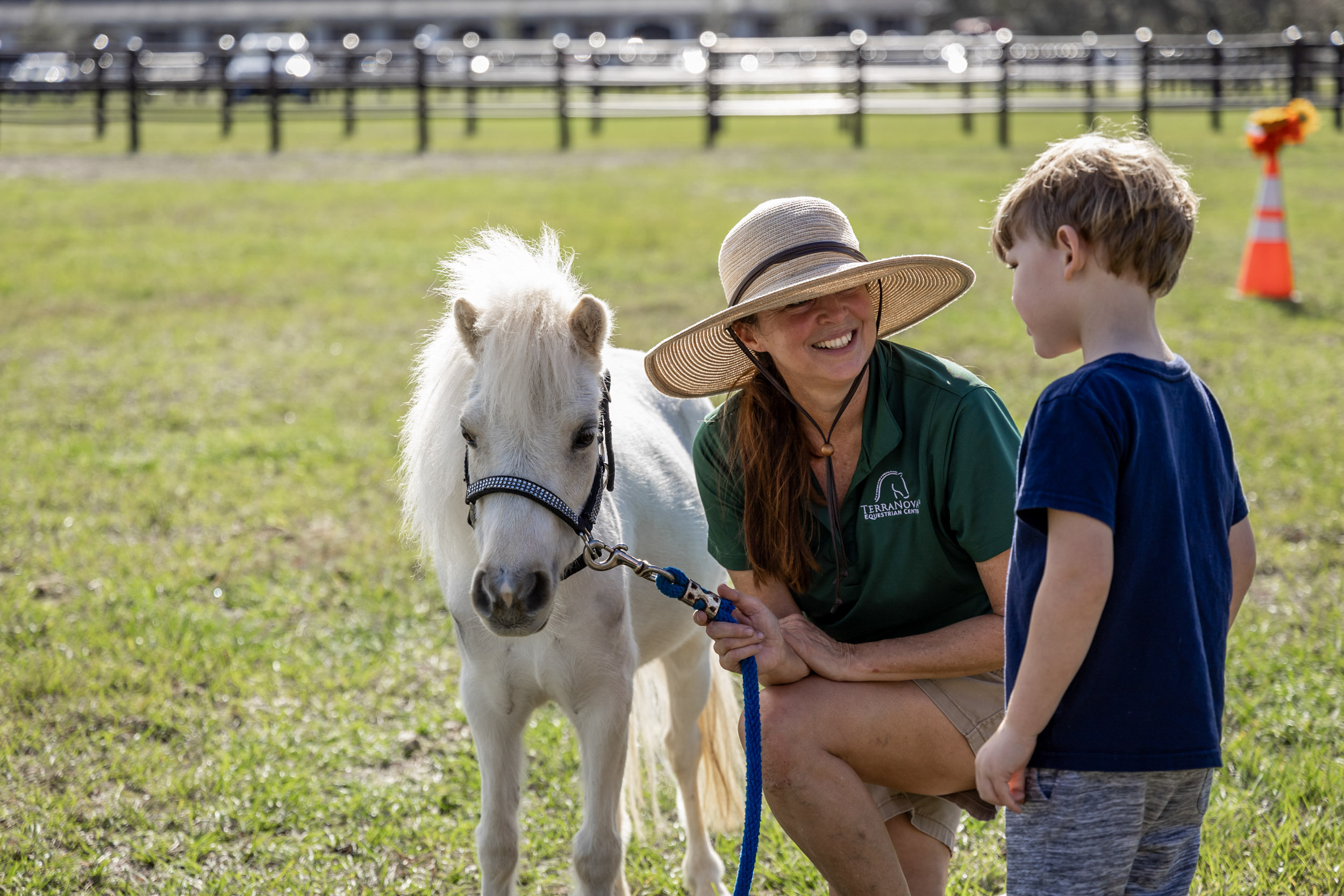Staff member in a sun hat kneels with a white miniature horse beside a young boy