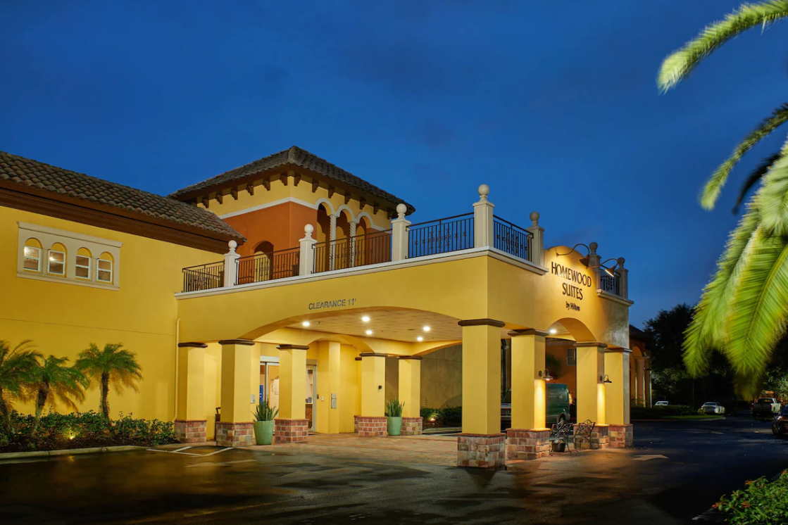 Yellow Homewood Suites hotel entrance lit at dusk with palms beside the drive