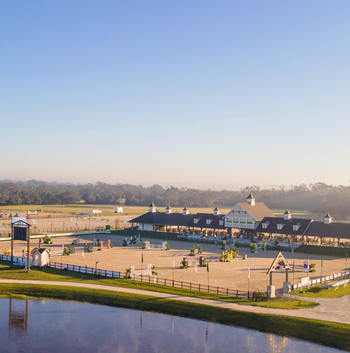 Aerial view of the TerraNova showgrounds at sunrise, with the covered arena pavilion, a sand jumping arena and a lake in the foreground.